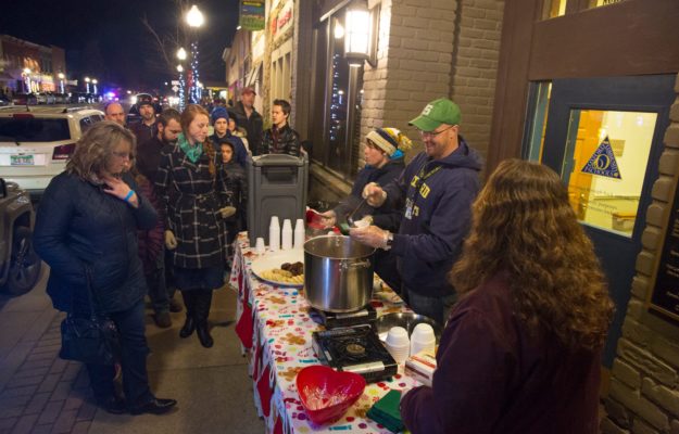 Oxford Community Shools volunteers for the Soup & Sweet Stroll serve soup and cookies to attendees outdoors on a cold December evening in Downtown Oxford