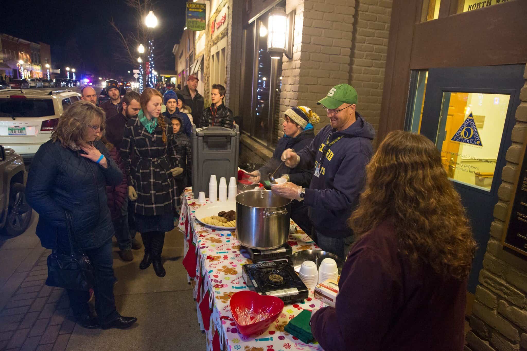 Oxford Community Shools volunteers for the Soup & Sweet Stroll serve soup and cookies to attendees outdoors on a cold December evening in Downtown Oxford